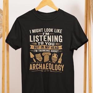 Könnte beinhalten: Ein schwarzes Kurzarm-T-Shirt mit beigefarbenem Text, der I MIGHT LOOK LIKE I'M LISTENING TO YOU BUT IN MY HEAD I'M THINKING ABOUT ARCHAEOLOGY lautet, ergänzt durch grafische Darstellungen einer Maurerkelle, Tonscherben, einer Säule, einer Vase und eines Pinsels.