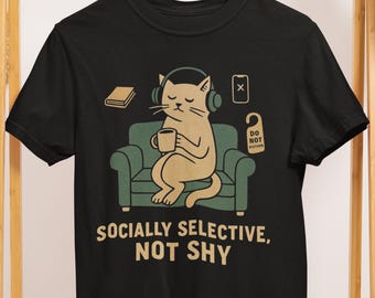 May include: Black t-shirt with a graphic of a cat wearing headphones, holding a mug, and sitting on a green couch. The text "SOCIALLY SELECTIVE, NOT SHY" is printed below the graphic. Additional graphics include a book, a phone, and a "DO NOT DISTURB" sign.