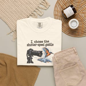 Könnte beinhalten: Ein cremefarbenes T-Shirt mit einer schwarzen Kamera auf einem Stativ und einem bunten Eisvogel, der ins Wasser eintaucht. Der Text "I chose the shutter-speed gamble" ist in Schwarz über der Grafik gedruckt. Das T-Shirt hat ein Comfort Colors Etikett.