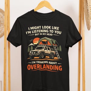 Könnte beinhalten: Schwarzes T-Shirt mit einer Grafik eines Geländewagens mit Dachzelt, Bäumen und einem Berg. Der Text lautet: „Ich mag so aussehen, als würde ich dir zuhören, aber in meinem Kopf – denke ich ans Overlanding.“