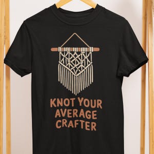 May include: Black t-shirt featuring a macrame design with the text "Knot Your Average Crafter" in brown. The macrame design is beige and brown, with a geometric pattern and fringe. The shirt is hanging on a wooden hanger.