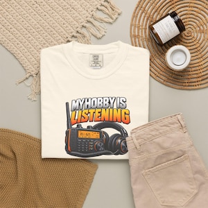 May include: A cream-coloured t-shirt featuring a graphic of a black two-way radio with an antenna and a pair of black over-ear headphones. The text above the radio reads "MY HOBBY IS LISTENING" in bold, white, and orange gradient lettering.