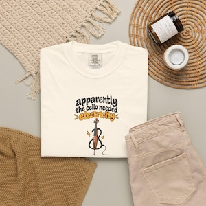 May include: A cream-colored t-shirt featuring the black text "apparently the cello needed" above the orange text "electricity" and a graphic of a brown cello with black lightning bolts and a black power cord extending from the base.