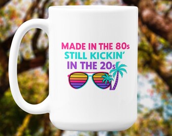 May include: White ceramic mug with a curved handle. The mug features the text "MADE IN THE 80s STILL KICKIN' IN THE 20s" in pink, teal, and blue. Below the text is a graphic of sunglasses and a palm tree in vibrant colors.