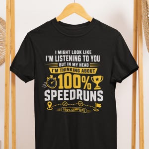 May include: A black short-sleeve t-shirt featuring white and yellow text that reads: I MIGHT LOOK LIKE I'M LISTENING TO YOU BUT IN MY HEAD I'M THINKING ABOUT 100% SPEEDRUNS. The design includes a stopwatch, a trophy, and a path with a flag icon.