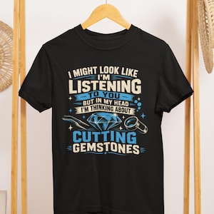 May include: A black short-sleeve t-shirt featuring the white and blue text I MIGHT LOOK LIKE I'M LISTENING TO YOU BUT IN MY HEAD I'M THINKING ABOUT CUTTING GEMSTONES, accompanied by a graphic illustration of a faceted diamond, tweezers, and a loupe.