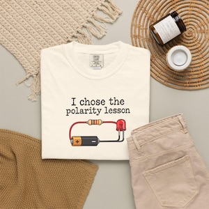 May include: A cream-colored t-shirt featuring the black text "I chose the polarity lesson" above a graphic of a simple electrical circuit. The circuit includes a battery, a resistor, and a red LED light connected by red and black wires.