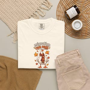 May include: A cream-colored short-sleeve t-shirt featuring a graphic of a white goose wearing a red and orange plaid scarf. The text above the goose reads "migration can wait" in orange and brown letters, surrounded by falling autumn leaves.