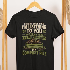 May include: A black short-sleeved t-shirt featuring the text I MIGHT LOOK LIKE I'M LISTENING TO YOU BUT IN MY HEAD I'M THINKING ABOUT MY COMPOST PILE in white and green lettering, accompanied by a graphic illustration of a wooden compost bin.