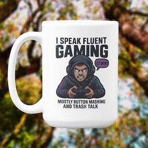 May include: White ceramic mug with a black handle featuring a gamer graphic. The graphic includes the text "I Speak Fluent Gaming" and "Mostly Button Mashing and Trash Talk." A cartoon character with headphones and a controller is also shown.