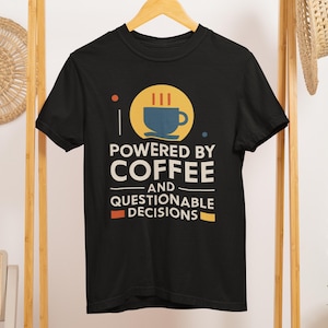 May include: Black t-shirt with the text "POWERED BY COFFEE AND QUESTIONABLE DECISIONS". The design includes a coffee cup graphic in a yellow circle. The shirt is hanging on a wooden hanger.