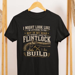 May include: A black short-sleeved t-shirt featuring a graphic print in tan and white. The text reads: I MIGHT LOOK LIKE I'M LISTENING TO YOU BUT IN MY HEAD I'M THINKING ABOUT MY FLINTLOCK BUILD. Below the text is an illustration of a flintlock pistol.