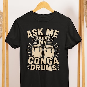 Op de afbeelding: Zwart t-shirt met de tekst "ASK ME ABOUT MY CONGA DRUMS" in een crèmekleurig lettertype. Het ontwerp toont twee conga drums met stralende lijnen, wat een muzikaal thema creëert.
