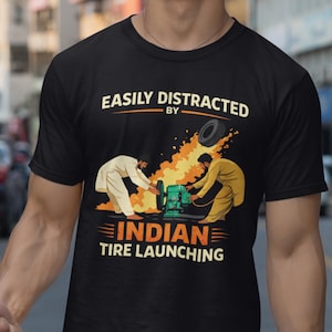 Puede incluir: Camiseta negra con el texto "EASILY DISTRACTED BY INDIAN TIRE LAUNCHING". El diseño muestra dos figuras con vestimenta tradicional operando una máquina de lanzamiento de neumáticos, con un neumático en el aire.