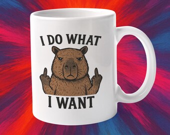 Op de afbeelding: Witte keramische mok met een bruine capibara-illustratie die de middelvinger opsteekt. De tekst "I DO WHAT I WANT" staat boven en onder de capibara. De mok heeft een gebogen handvat en een glanzende afwerking.