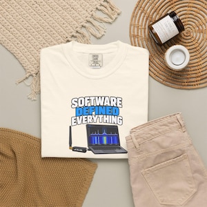 Könnte beinhalten: Ein cremefarbenes T-Shirt mit dem Text "SOFTWARE DEFINED EVERYTHING" in schwarzer und blauer Schrift. Unter dem Text befindet sich eine Grafik eines Laptops, der einen Funkfrequenz-Spektrumanalysator zeigt, verbunden mit einem RTL-SDR USB-Stick.