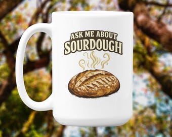Könnte beinhalten: Weißer Keramikbecher mit Henkel, mit dem Text "ASK ME ABOUT SOURDOUGH" über einer Illustration eines braunen Brotlaibs. Dampf steigt vom Brot auf. Der Hintergrund ist verschwommen, mit Grün- und Brauntönen.