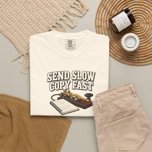 Op de afbeelding: Een crèmekleurig T-shirt met een afbeelding van een vintage telegraafsleutel en een spiraalblok. De tekst boven de illustratie luidt "SEND SLOW COPY FAST" in dikke, donkere letters. Het T-shirt is voorzien van een Comfort Colors label.