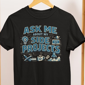 以下が含まれることがあります： 「ASK ME ABOUT MY SIDE PROJECTS」と青い文字で書かれた黒いTシャツ。デザインには、電球、ラップトップ、ロケット、コーヒーカップ、その他のテクノロジー関連のグラフィックが含まれています。開発者やテクノロジー愛好家のための楽しいTシャツです。