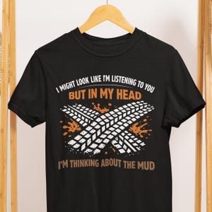 Könnte beinhalten: Schwarzes T-Shirt mit dem Text "I MIGHT LOOK LIKE I'M LISTENING TO YOU BUT IN MY HEAD I'M THINKING ABOUT THE MUD." Das Shirt zeigt ein Reifenabdruck-Design mit orangefarbenen Schlammspritzern.