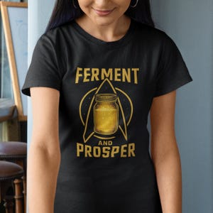 Puede incluir: Camiseta negra con un diseño dorado. El texto "FERMENT AND PROSPER" rodea un tarro con líquido dorado. La camiseta es de manga corta.