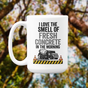 Op de afbeelding: Witte keramische mok met de tekst "I LOVE THE SMELL OF FRESH CONCRETE IN THE MORNING". De mok heeft een afbeelding van een betonmixer, oranje verkeerskegels en een geel-zwarte gestreepte rand.