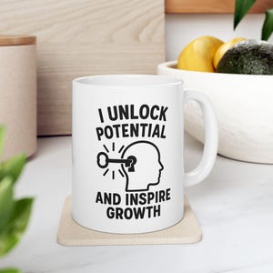 Può includere: Tazza in ceramica bianca con la scritta "I UNLOCK POTENTIAL AND INSPIRE GROWTH" in nero. Il design include una chiave che sblocca una silhouette di testa. La tazza è su un sottobicchiere beige.