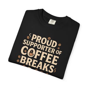 May include: Black t-shirt with the text "PROUD SUPPORTER OF COFFEE BREAKS" in a light brown color. The design includes coffee beans and a coffee cup icon. The shirt is made of a soft material.