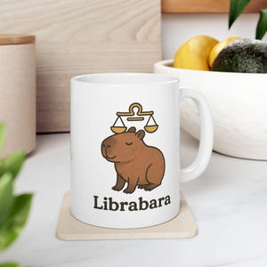 Könnte beinhalten: Weiße Keramiktasse mit einer braunen Capybara-Illustration und einem gelben Waage-Symbol. Der Text "Librabara" ist auf die Tasse gedruckt.