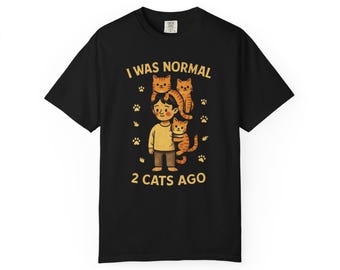 May include: Black t-shirt with a graphic featuring a cartoon person with three orange cats and paw prints. The text reads "I WAS NORMAL 2 CATS AGO" in a playful font. A fun, cat-themed apparel item.