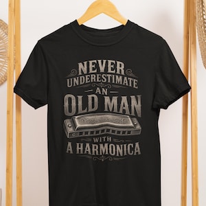 May include: Black t-shirt with the text "NEVER UNDERESTIMATE AN OLD MAN WITH A HARMONICA" in a vintage style. The design includes an illustration of a harmonica. The shirt is hanging on a wooden hanger.