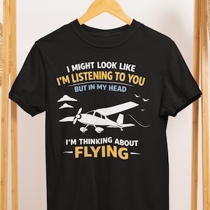 May include: Black t-shirt with a white graphic of a small airplane and the text "I MIGHT LOOK LIKE I'M LISTENING TO YOU BUT IN MY HEAD I'M THINKING ABOUT -FLYING-". The shirt is hanging on a wooden hanger.