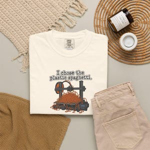 Peut inclure: Un t-shirt couleur crème avec un motif représentant une imprimante 3D et un amas de filament orange emmêlé ressemblant à des spaghettis. Le texte au-dessus de l'imprimante indique : "I chose the plastic spaghetti." Étiquette Comfort Colors.