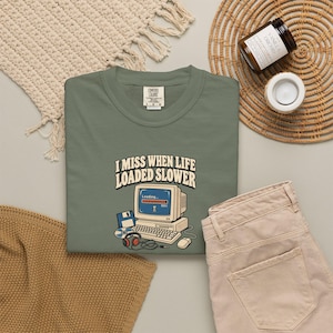 May include: An olive green t-shirt featuring a graphic of a vintage computer with the text "I MISS WHEN LIFE LOADED SLOWER" above it. The design includes a floppy disk, a compact disc, headphones, and a computer mouse surrounding the monitor.