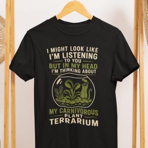 May include: A black short-sleeved t-shirt featuring a graphic of a glass terrarium containing various carnivorous plants. The text reads: I MIGHT LOOK LIKE I'M LISTENING TO YOU BUT IN MY HEAD I'M THINKING ABOUT MY CARNIVOROUS PLANT TERRARIUM.