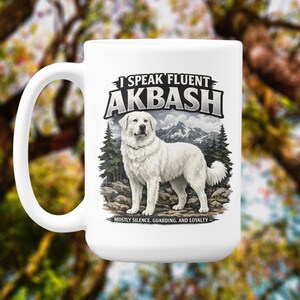 Puede incluir: Taza de cerámica blanca con un gráfico de un perro Akbash blanco frente a un paisaje montañoso. El texto en la taza dice "I Speak Fluent Akbash" y "Mostly Silence, Guarding, and Loyalty".