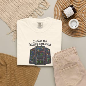 May include: A cream-coloured t-shirt featuring a graphic of three black server racks with colourful blinking lights and the text "I chose the blinking-light oracle." The shirt has a Comfort Colors brand label at the inner neckline showing size L/G.