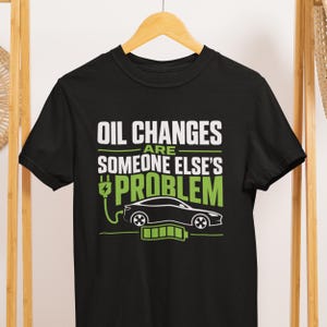 May include: A black short-sleeve t-shirt featuring white and green text that reads OIL CHANGES ARE SOMEONE ELSE'S PROBLEM. The graphic includes a white outline of an electric car, a green power plug icon, and a green battery charge indicator.