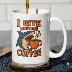 Puede incluir: Taza de cerámica blanca con el texto "I BITE BEFORE COFFEE" en letras naranjas. Un tiburón de dibujos animados con la boca abierta y un gráfico de taza de café también aparecen en la taza. Se está vertiendo café en la taza.