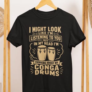 Op de afbeelding: Zwart t-shirt met een humoristische afbeelding. De tekst luidt: "I MIGHT LOOK LIKE I'M LISTENING TO YOU BUT IN MY HEAD I'M THINKING ABOUT MY CONGA DRUMS." Het ontwerp bevat illustraties van conga drums.