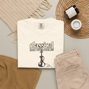May include: A cream-colored short-sleeve t-shirt featuring the text "classical but with cables" in a dark brown font, accompanied by a graphic of a brown electric violin with a black cable extending from the base of the instrument.