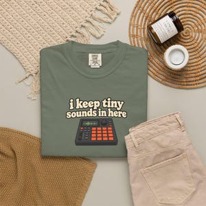 May include: An olive green t-shirt featuring the text "i keep tiny sounds in here" in a cream-colored font above a graphic illustration of a black music sampler device with orange square buttons and a small screen displaying a green sound wave.