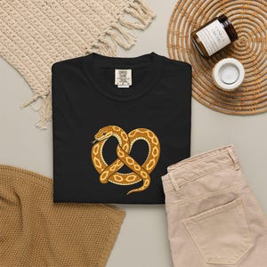 Può includere: Una t-shirt nera con la grafica di un serpente marrone e beige attorcigliato a forma di pretzel. Il serpente presenta un disegno a scaglie e una piccola lingua visibile. La maglietta ha un'etichetta del marchio Comfort Colors sul collo.