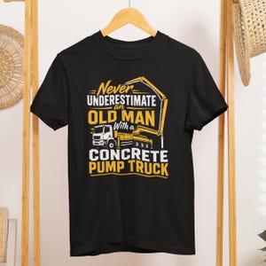 May include: A black short-sleeved t-shirt featuring a yellow and white graphic of a concrete pump truck. The text reads: Never underestimate an old man with a concrete pump truck. The design is centered on the chest of the garment.