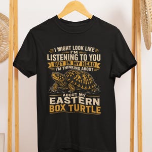 Könnte beinhalten: Ein schwarzes Kurzarm-T-Shirt mit einer Grafik einer östlichen Dosenschildkröte. Der Text lautet: I MIGHT LOOK LIKE I'M LISTENING TO YOU BUT IN MY HEAD I'M THINKING ABOUT MY EASTERN BOX TURTLE. Das Design verwendet gelbe und weiße Schriftzüge.