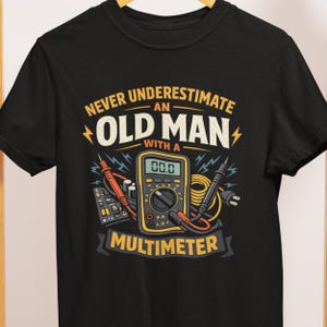 May include: Black t-shirt with a graphic featuring a multimeter and the text "Never Underestimate an Old Man with a Multimeter." The design includes lightning bolts and a yellow and black color scheme.
