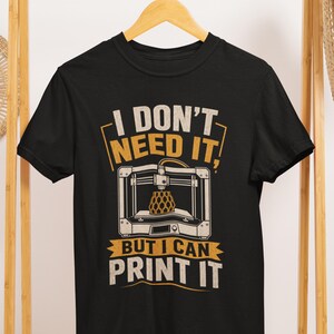 May include: A black short-sleeved t-shirt featuring a graphic of a 3D printer creating a geometric object. The text "I DON'T NEED IT, BUT I CAN PRINT IT" is printed in a distressed, off-white and yellow font above and below the printer illustration.