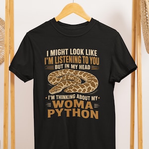 May include: A black short-sleeved t-shirt featuring a graphic of a brown and tan patterned snake. The text reads: I MIGHT LOOK LIKE I'M LISTENING TO YOU BUT IN MY HEAD I'M THINKING ABOUT MY WOMA PYTHON. The design is printed in tan and white ink.