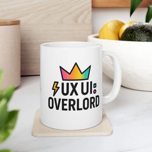 以下が含まれることがあります： 黒と虹色の王冠のグラフィックと「UX UI OVERLORD」というテキストが書かれた白いセラミックマグカップです。
