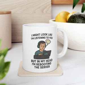 May include: White ceramic mug with a black outline illustration of a person wearing headphones and looking at a computer screen. The text on the mug reads "I might look like I'm listening to you, but in my head I'm rebooting the server."